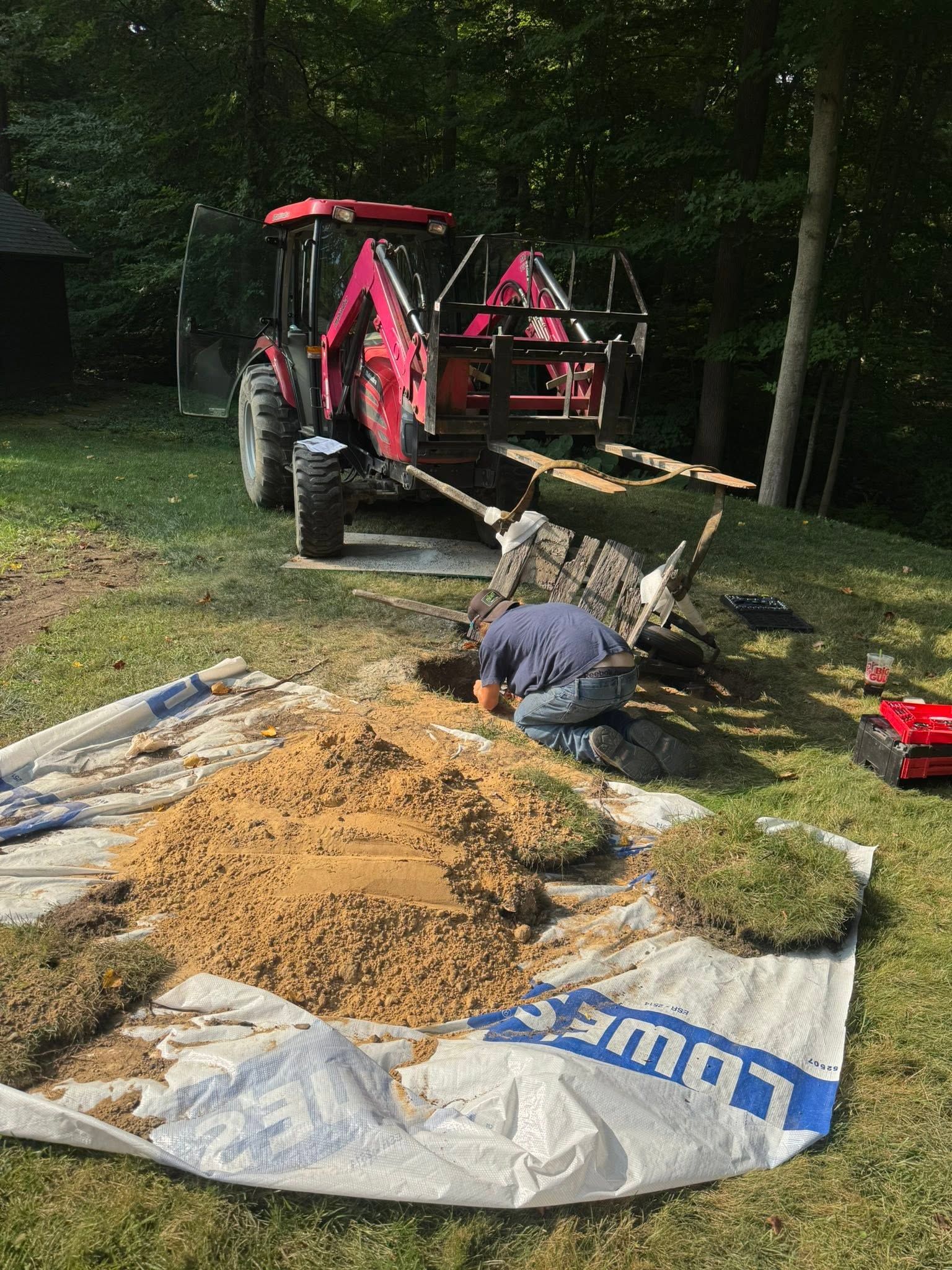 Tractor by a gravel pile with a worker in a wooded yard, handling a shovel near a tarp-covered area