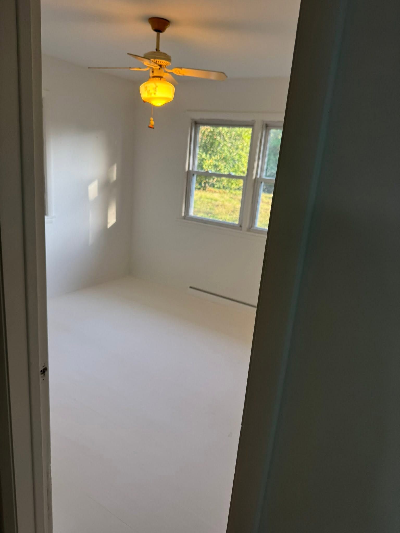 Empty white bedroom with a ceiling fan and a window letting in sunlight.