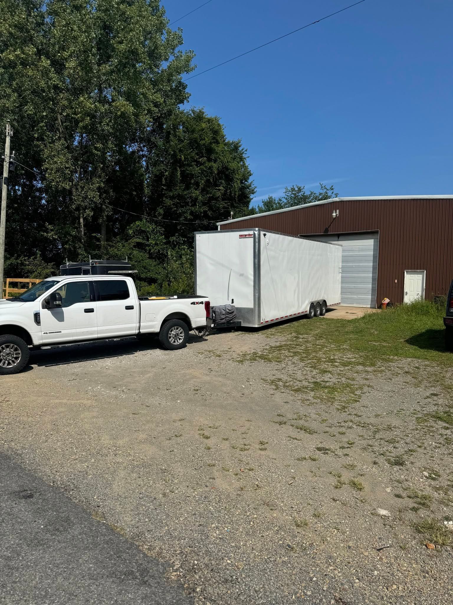 White pickup truck beside a white box trailer and brown building on a gravel lot under a clear blue sky.
