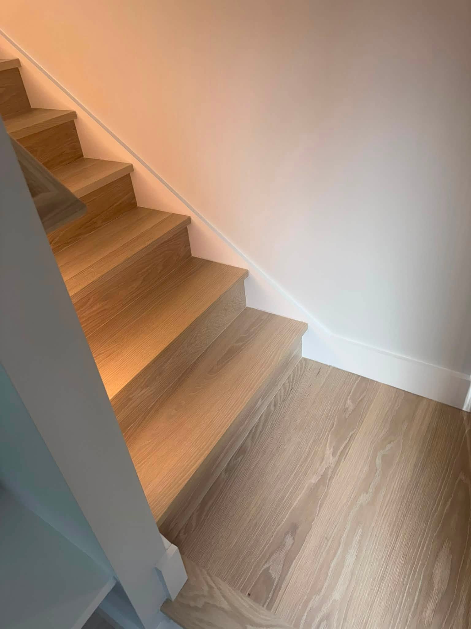 Interior staircase with light wood steps and matching wood flooring beside a white wall.