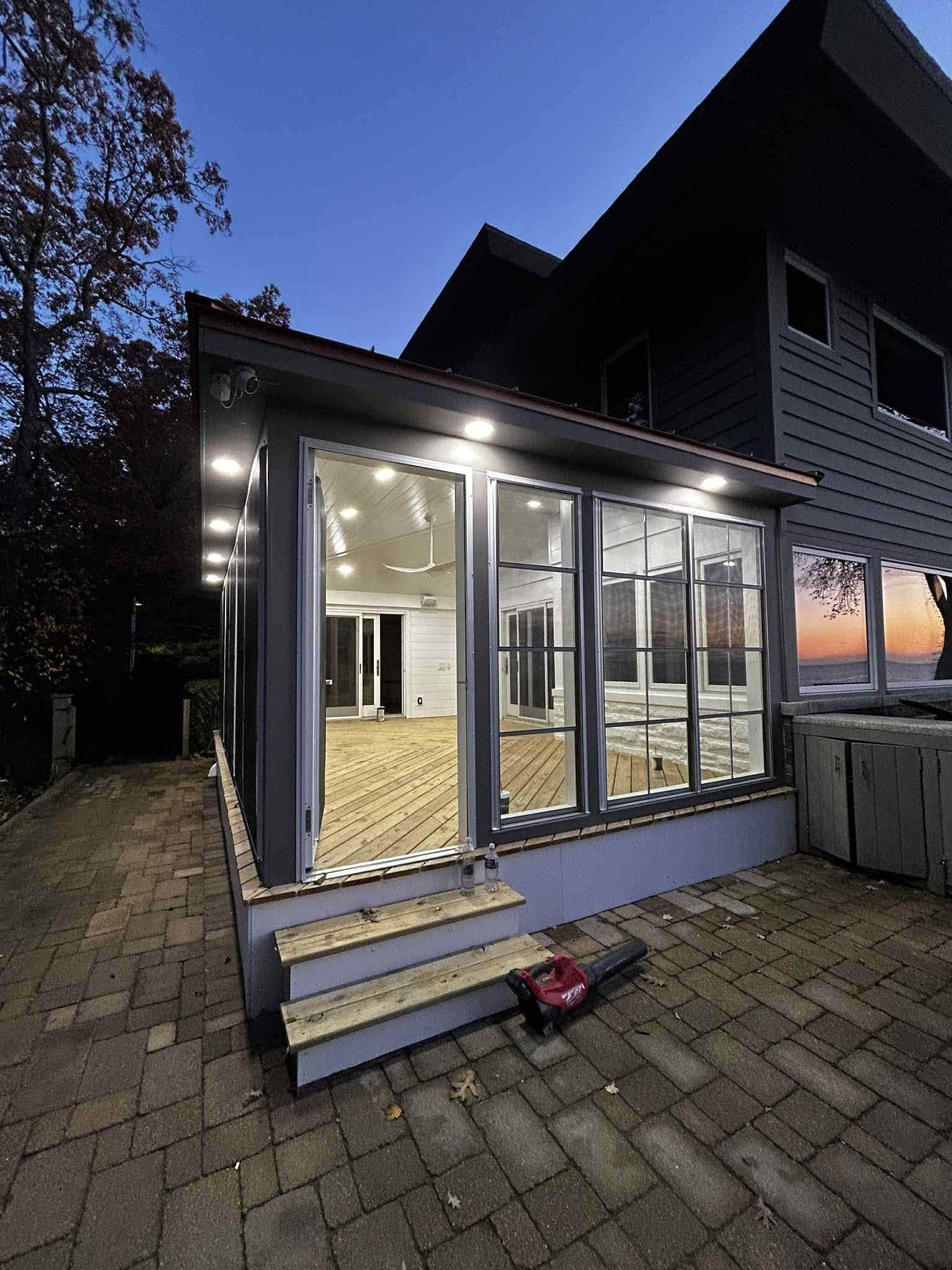 Modern glass sunroom addition with lit interior and steps at dusk, beside a brick patio and dark house