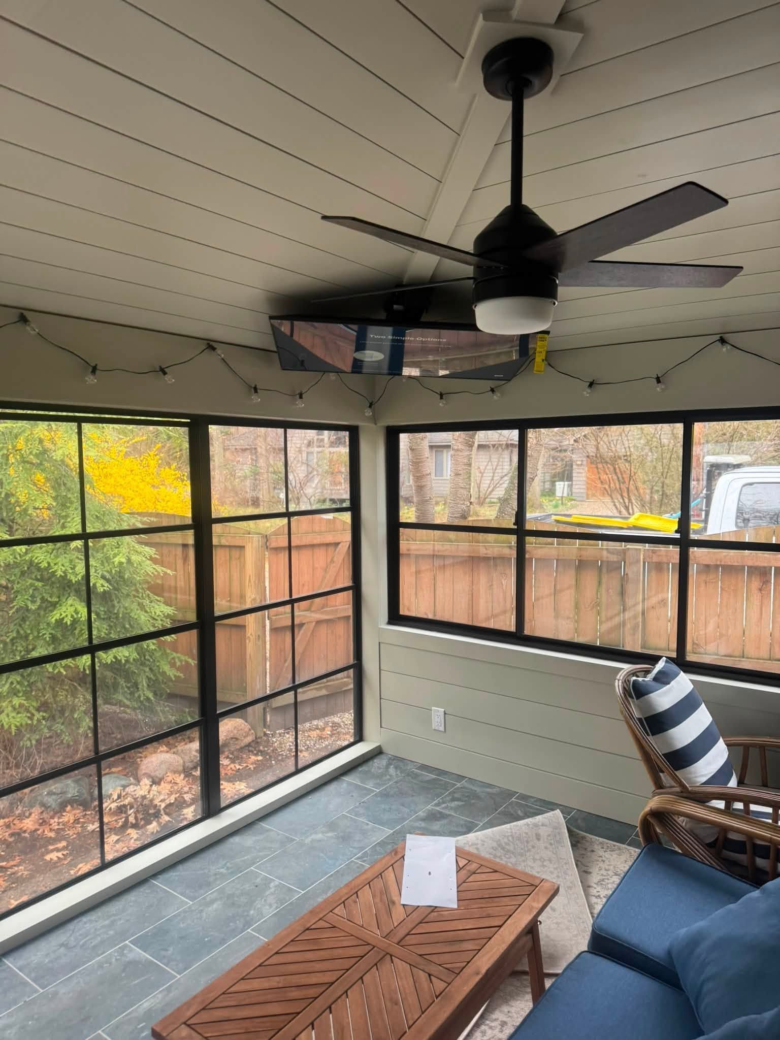 Screened porch with ceiling fan, blue chair, wooden table, and autumn trees outside