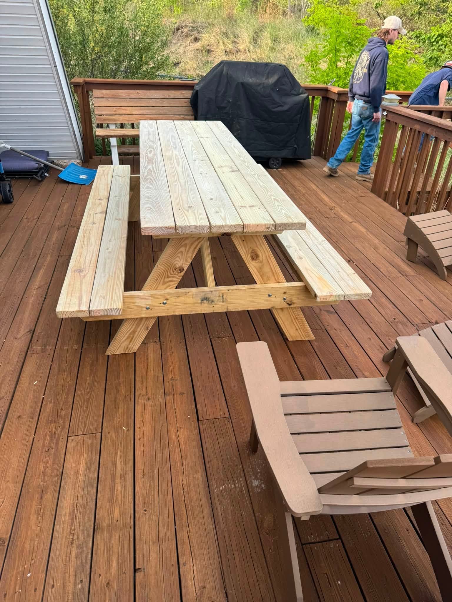 Wooden picnic table on a deck beside a covered grill, with a person standing in the background.