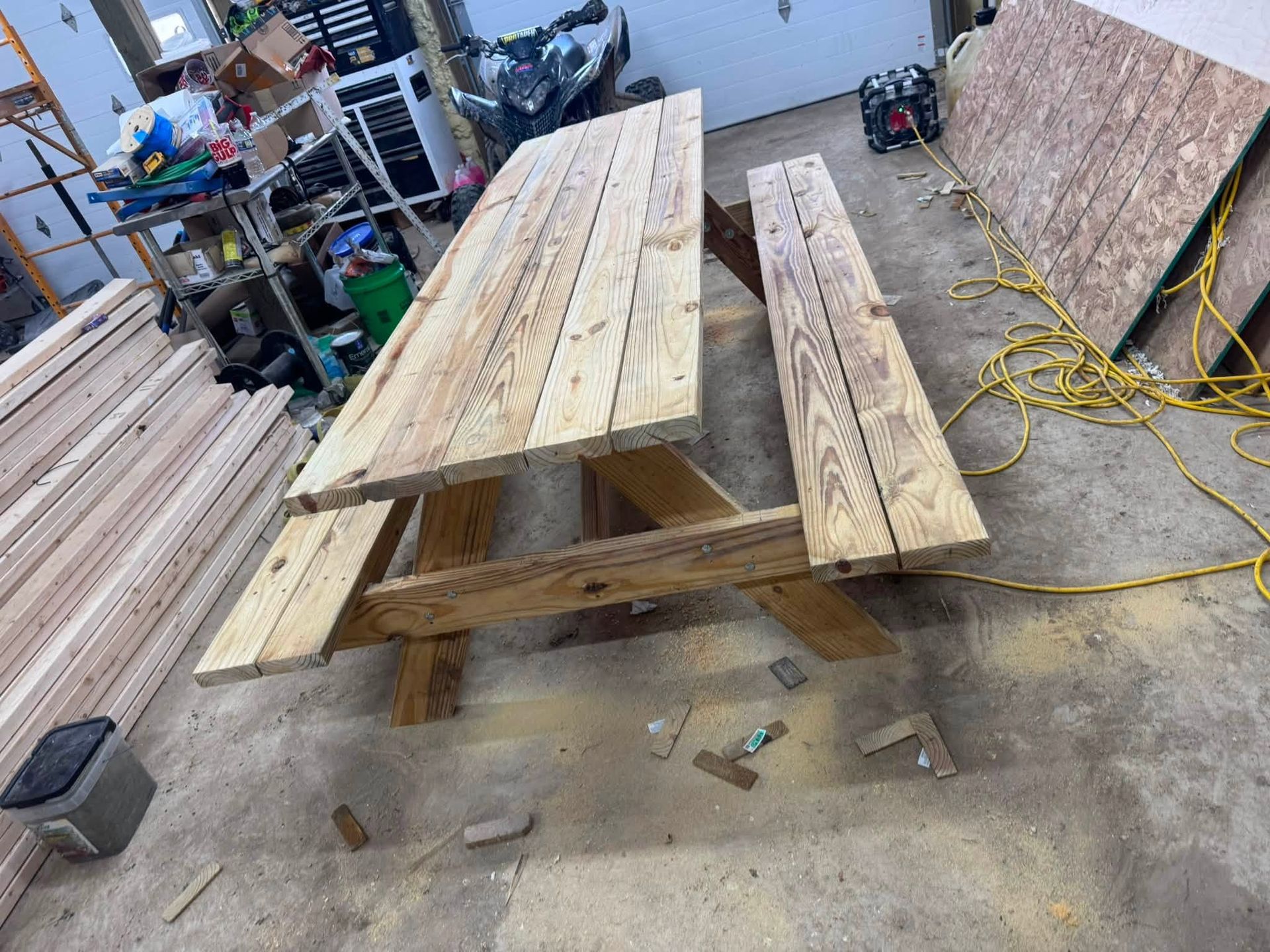 Wooden picnic table on a workshop floor with lumber, tools, and extension cords nearby