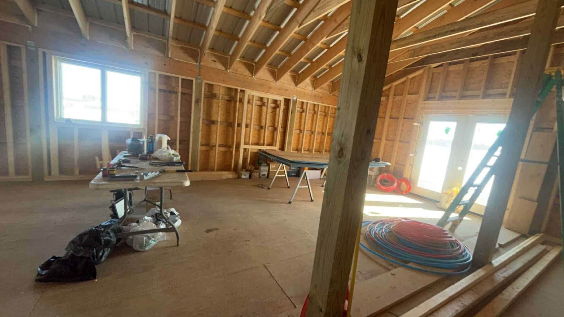 Unfinished attic room with exposed wooden rafters, work table, ladder, and tools scattered on the floor.