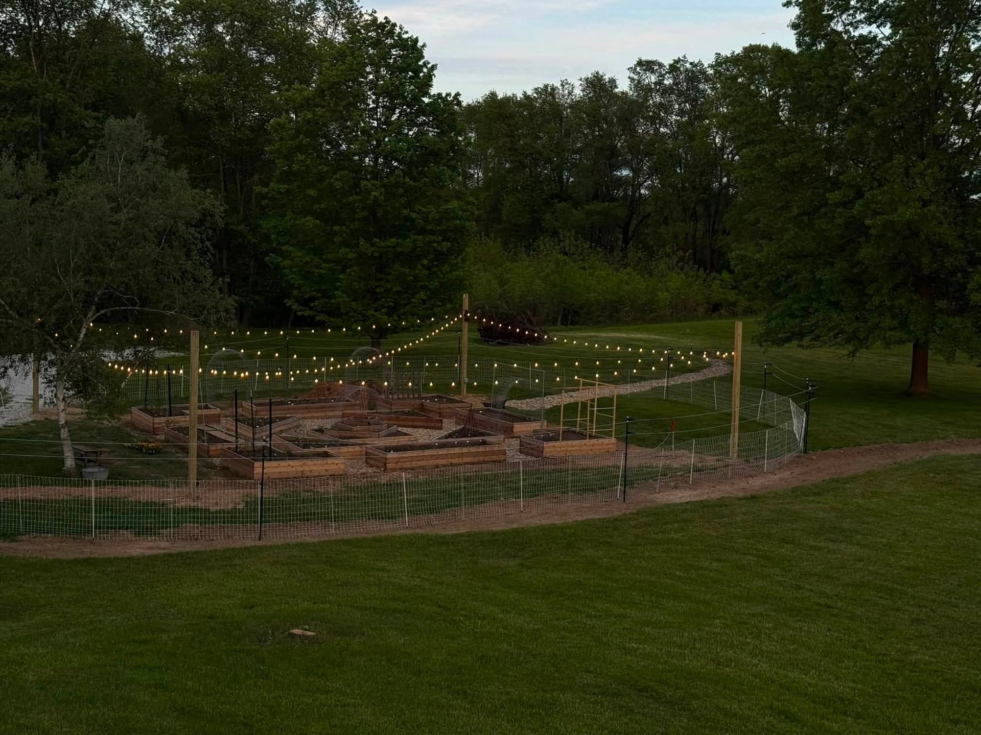 Small fenced garden with string lights and brick paths in a grassy park at dusk