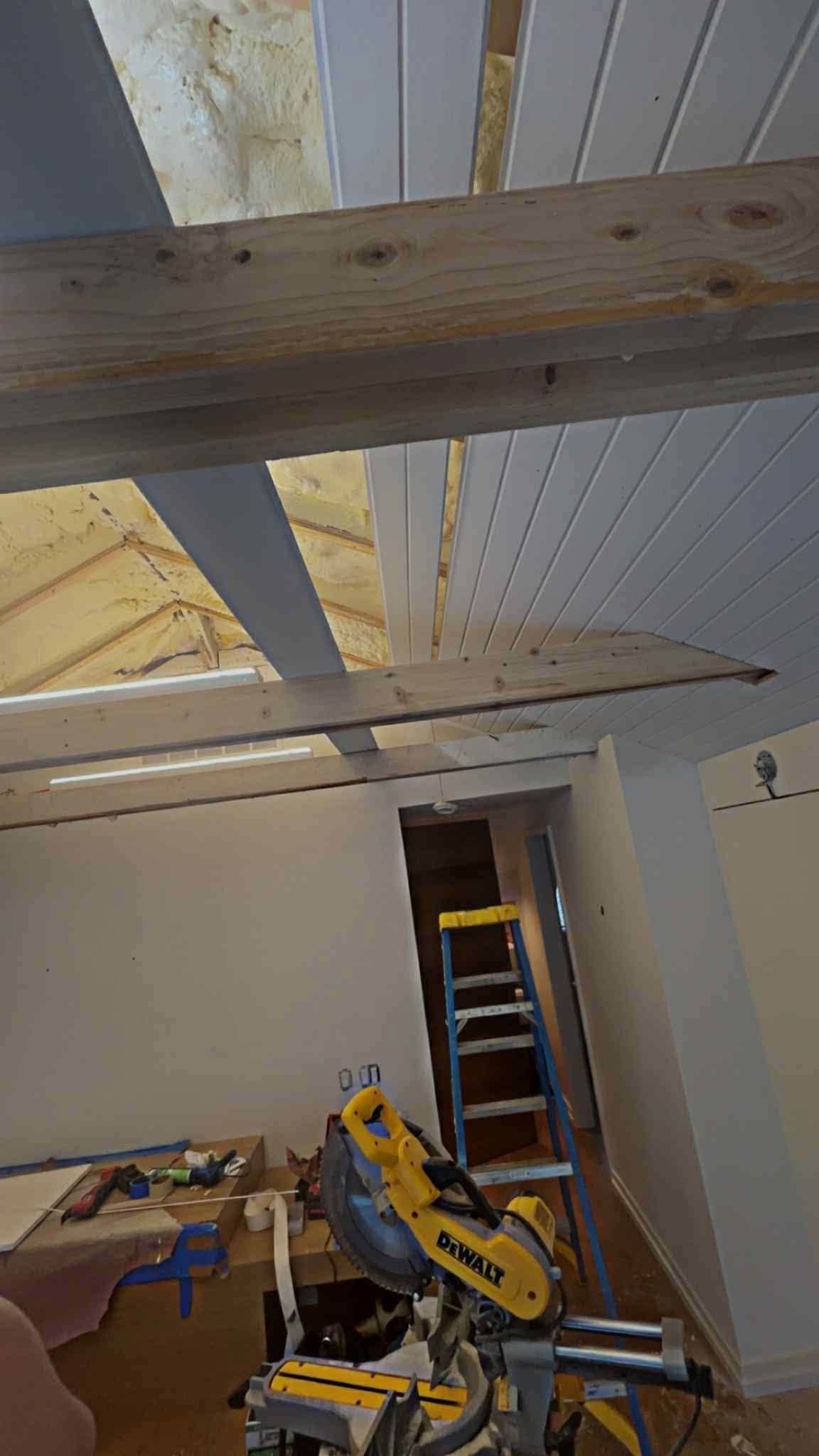 Interior room with slanted white ceiling, exposed wooden beams, and a yellow miter saw on a stand.