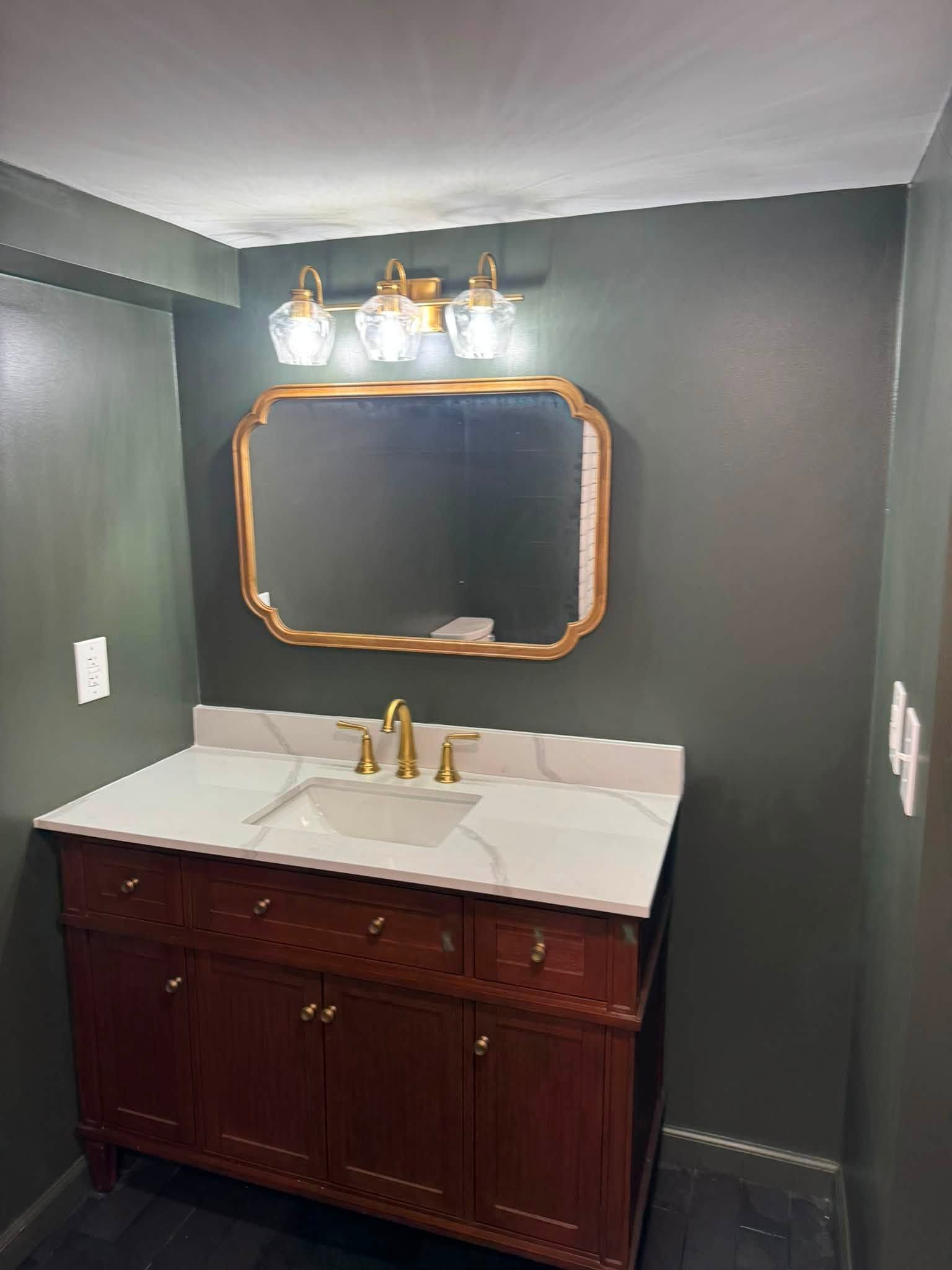 Bathroom vanity with dark wood cabinet, white sink, gold faucet, and ornate mirror on gray walls