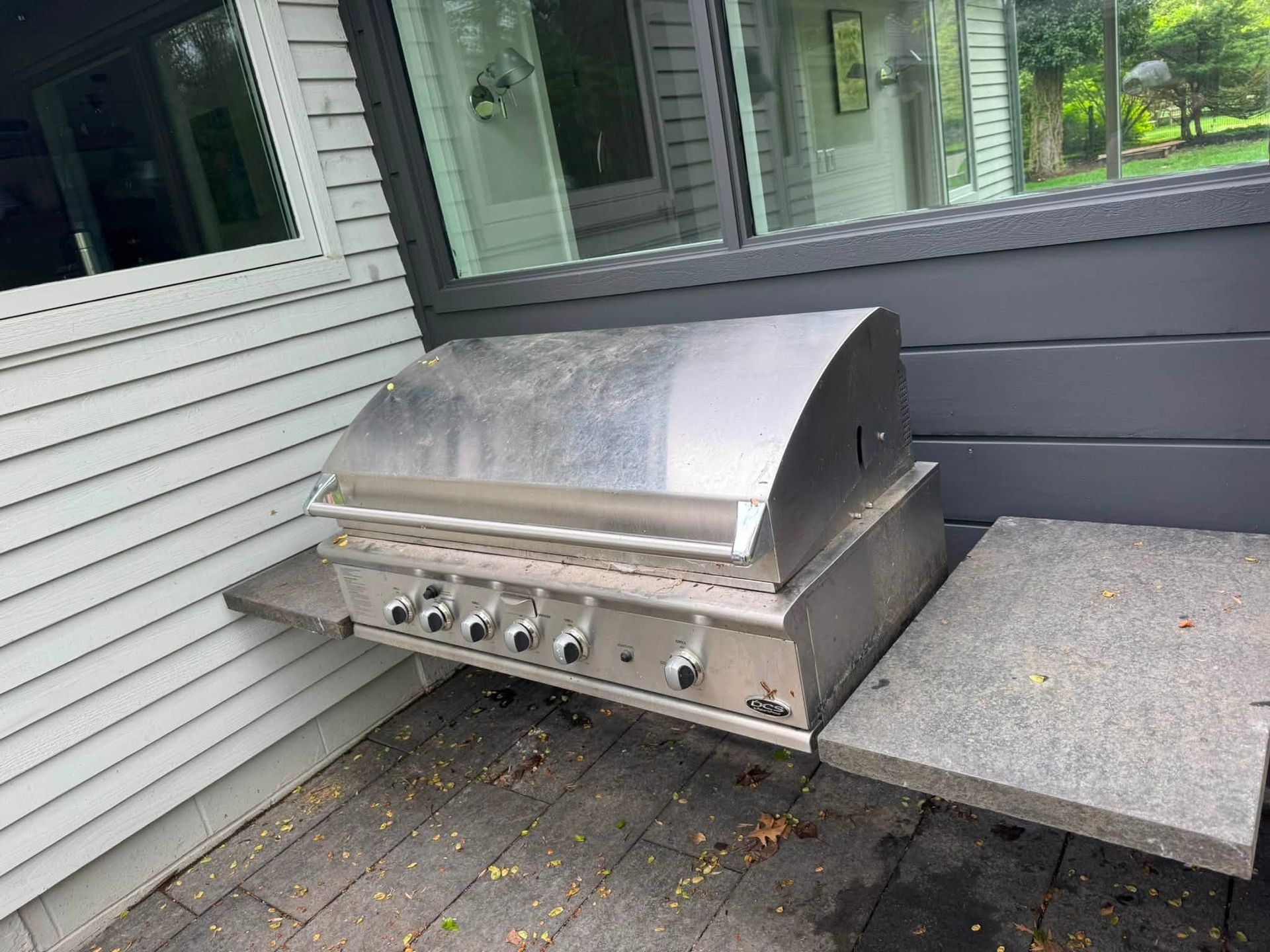 Stainless steel gas grill on a patio beside a house wall and window