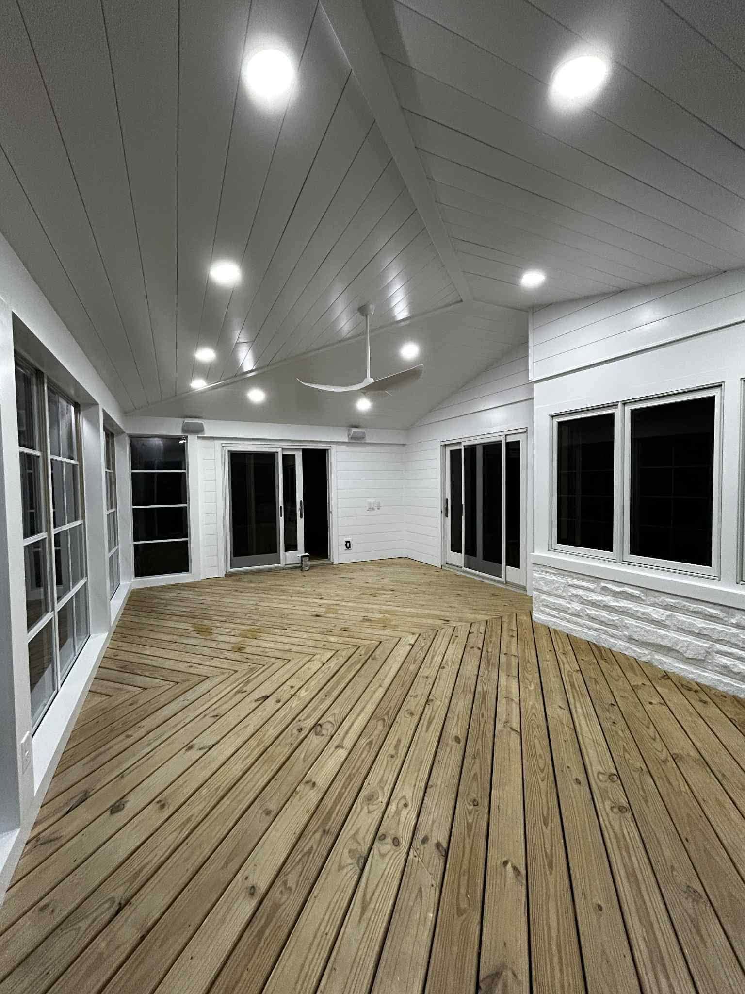 Empty enclosed porch with white walls, recessed lights, large windows, and a wood-plank floor under construction