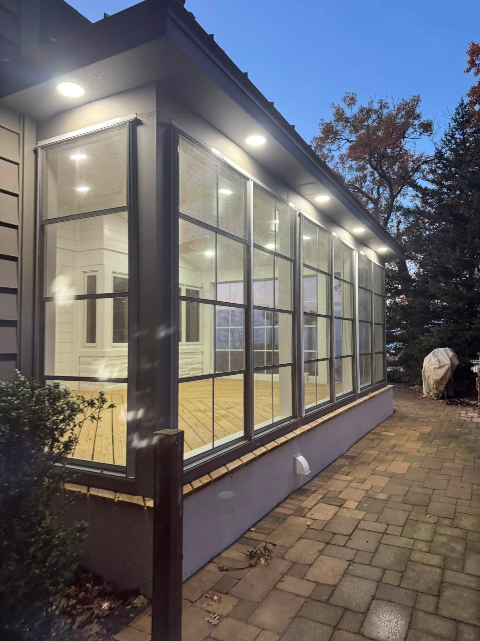 Glass-walled sunroom at dusk with warm interior lights beside a stone patio and pathway