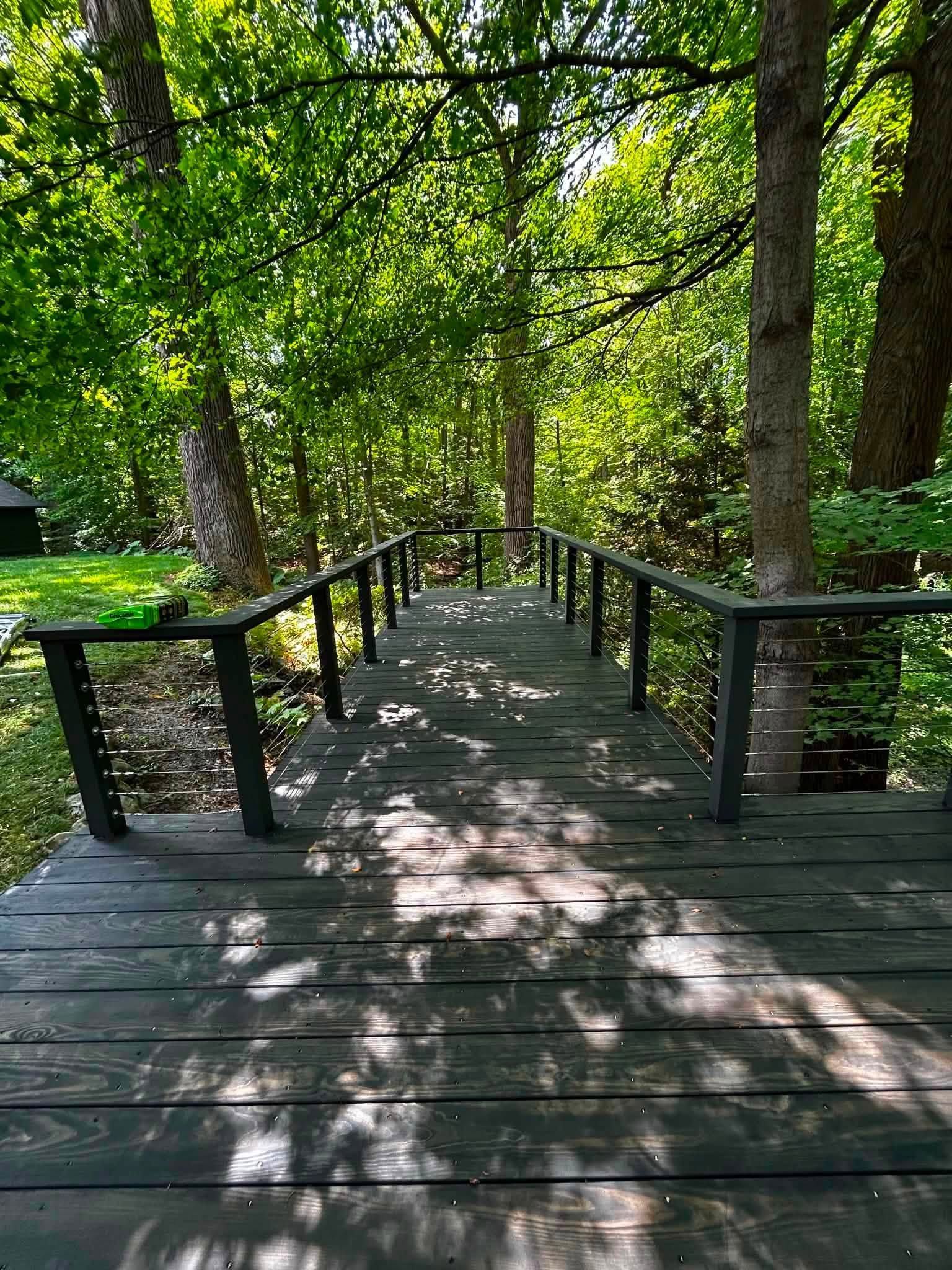 Wooden boardwalk through a leafy green forest, with dappled sunlight and black railings.