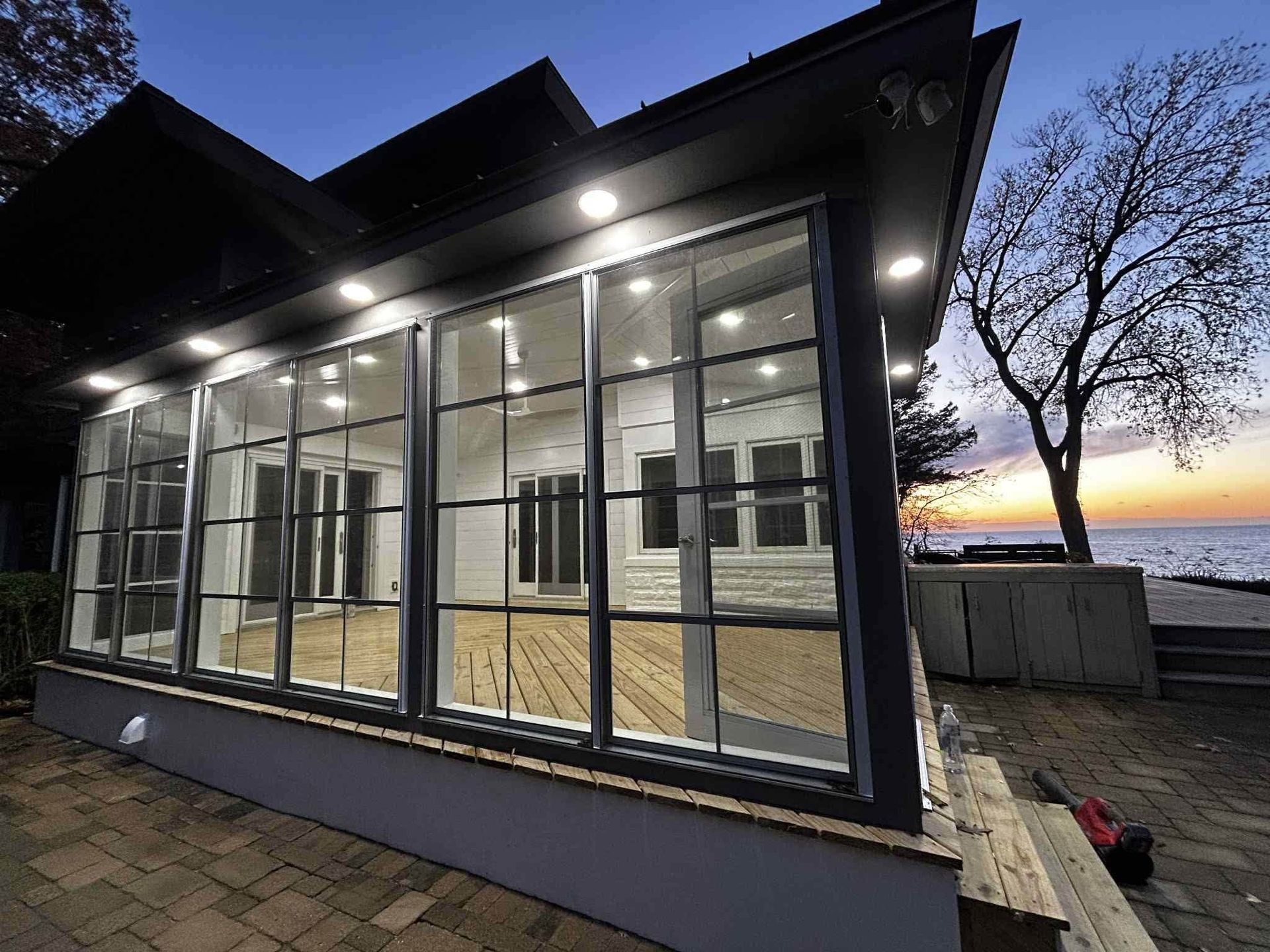 Modern glass-enclosed extension beside a house at dusk, with lit interior and waterfront view.