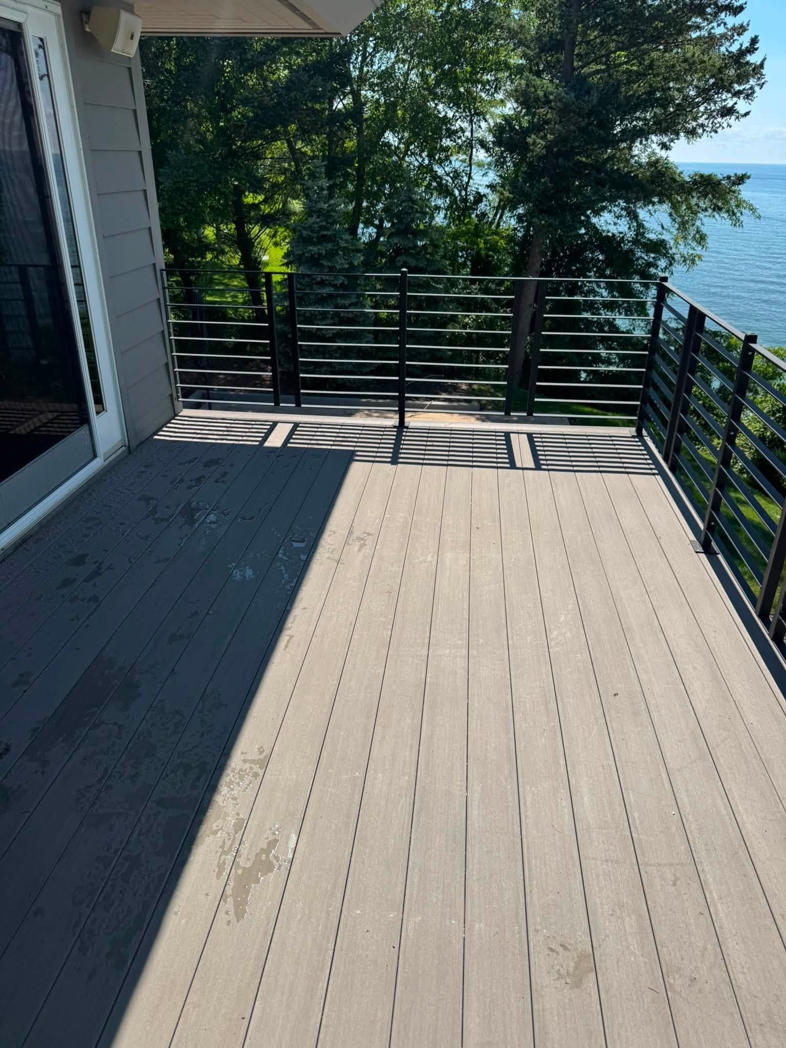 Sunlit outdoor deck with gray flooring and black railing overlooking trees and water