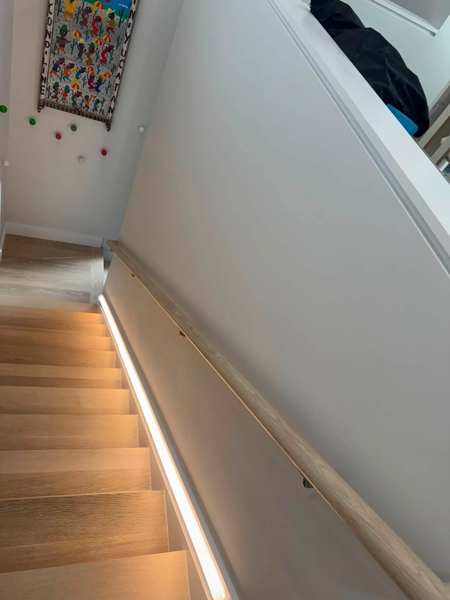 Staircase with wooden steps, white walls, and a lit handrail in a narrow indoor hallway