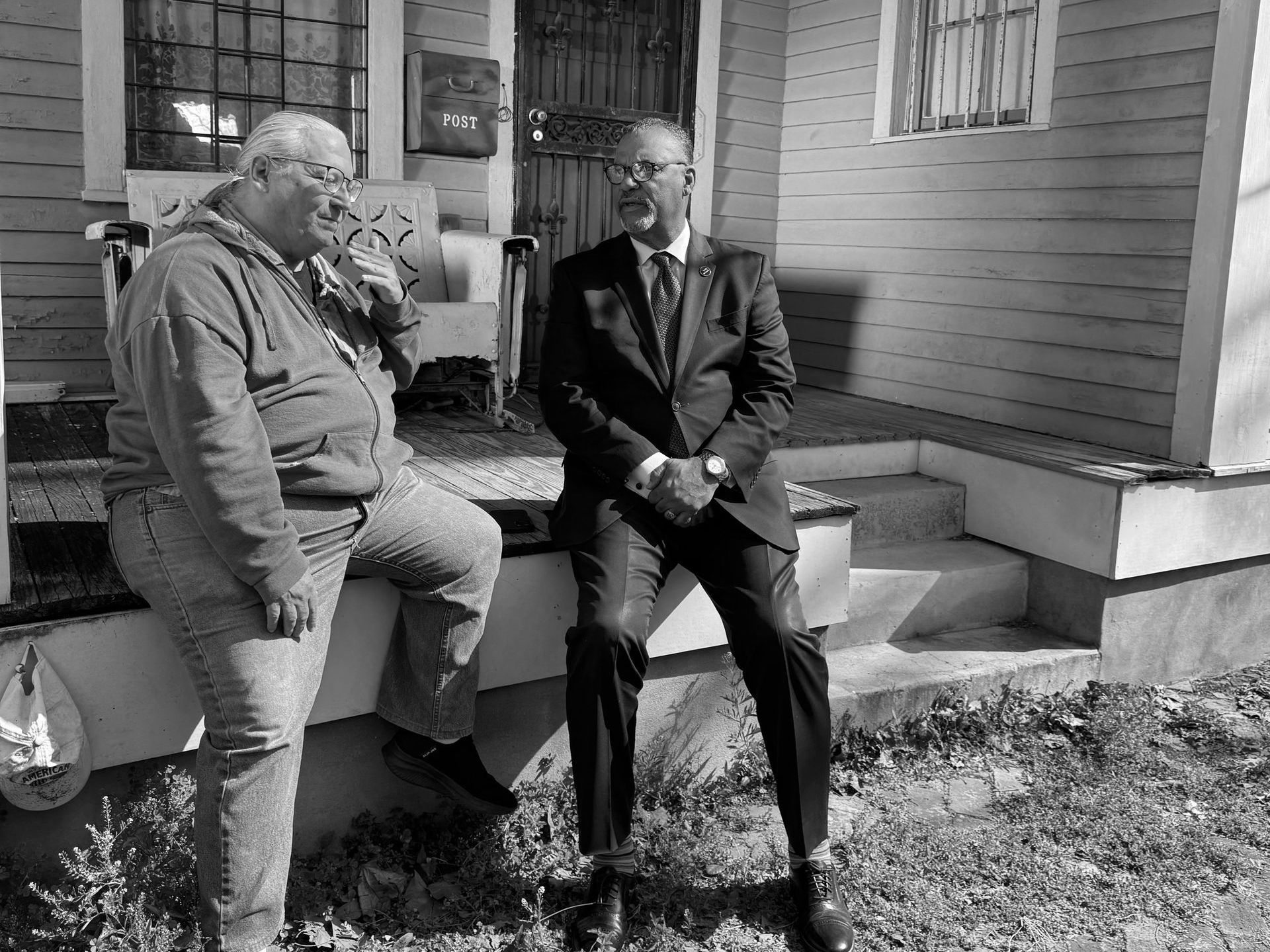 Oliver Thomas is sitting with a man on the porch of a house talking to each other.