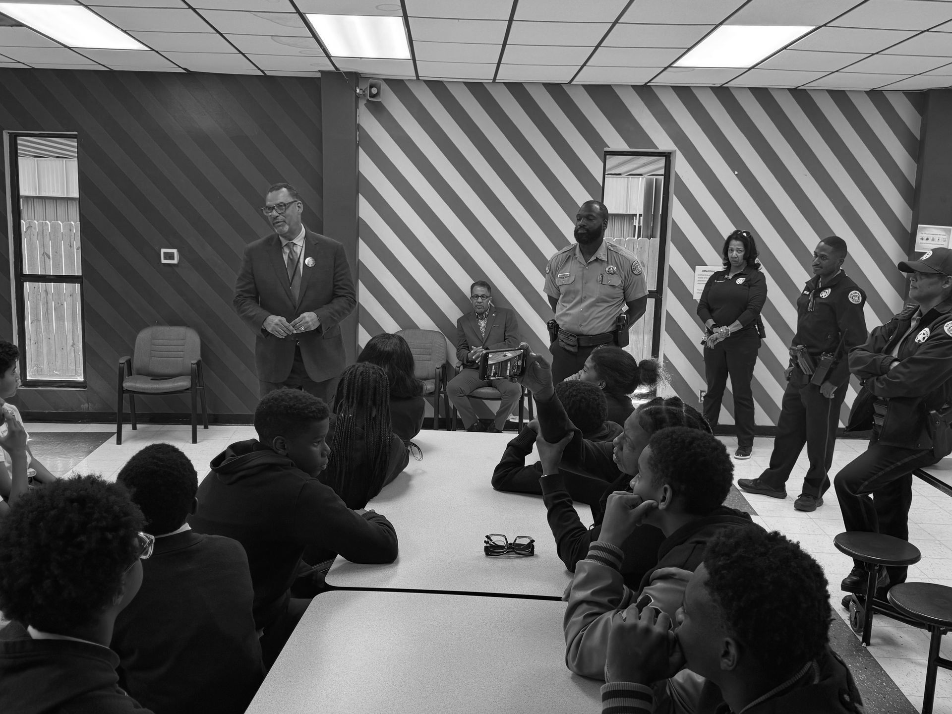 A black and white photo of a group of people sitting around a table with Oliver Thomas standing.