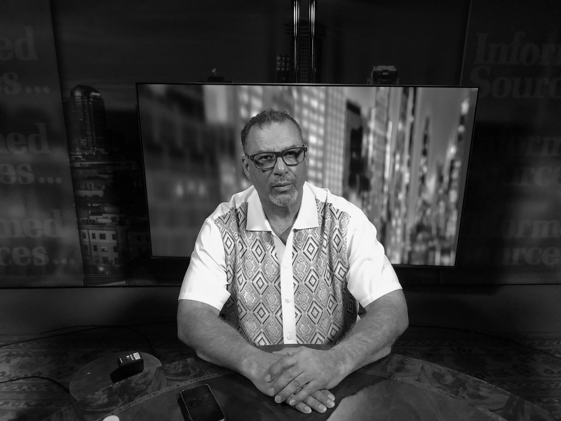 A black and white photo of Oliver Thomas sitting in front of a screen for an interview on national news.