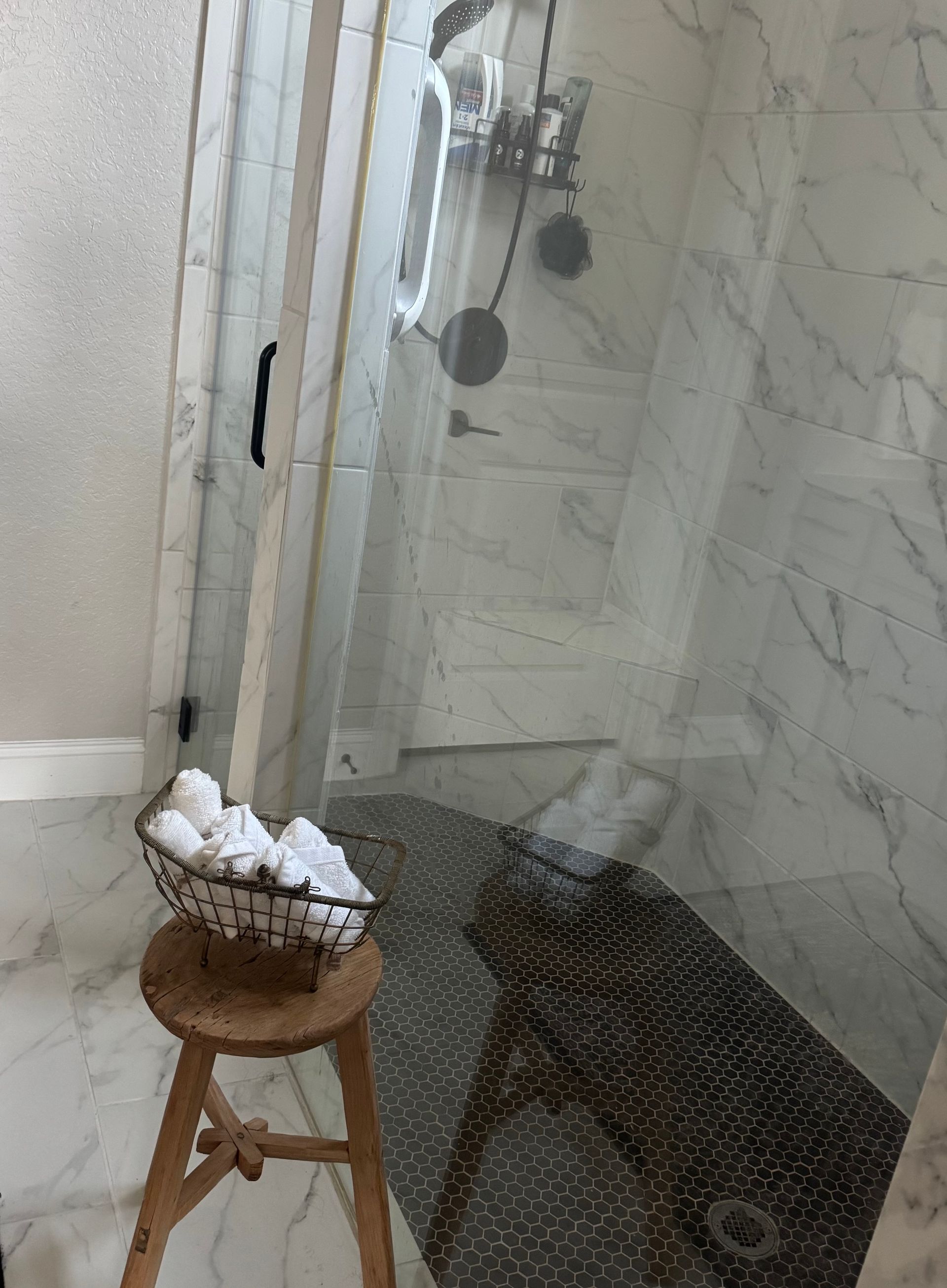 A basket of towels is sitting on a wooden stool in a bathroom next to a walk in shower.
