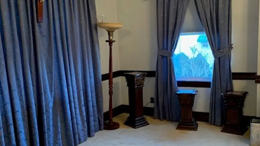 Room with blue curtains, dark pedestals, a floor lamp, and a window with outside foliage.