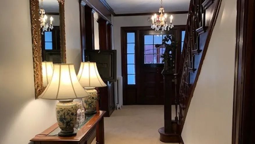 Hallway with antique lamps, mirror, and staircase with dark wood. A chandelier hangs above the doorway.