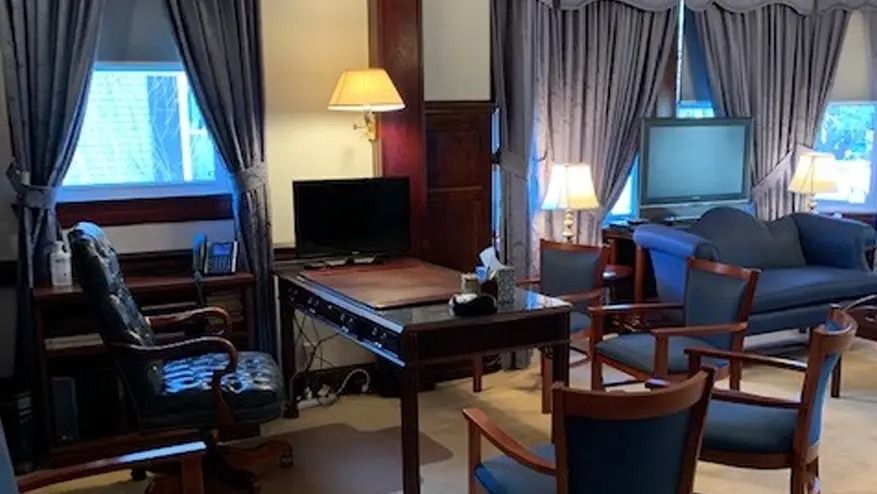 Office with desk, chairs, sofa, windows with drapes, lamps, and a television.