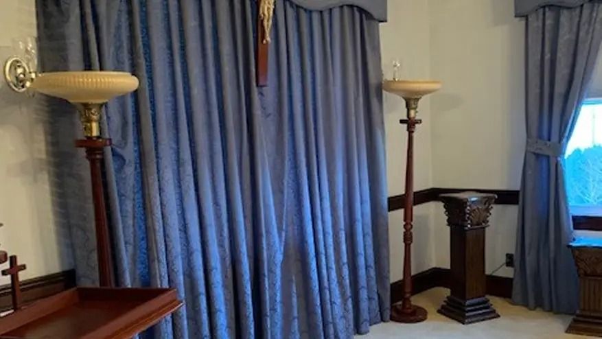 Two tall lamps, blue curtains, and a dark wood stand against a white wall in a room.