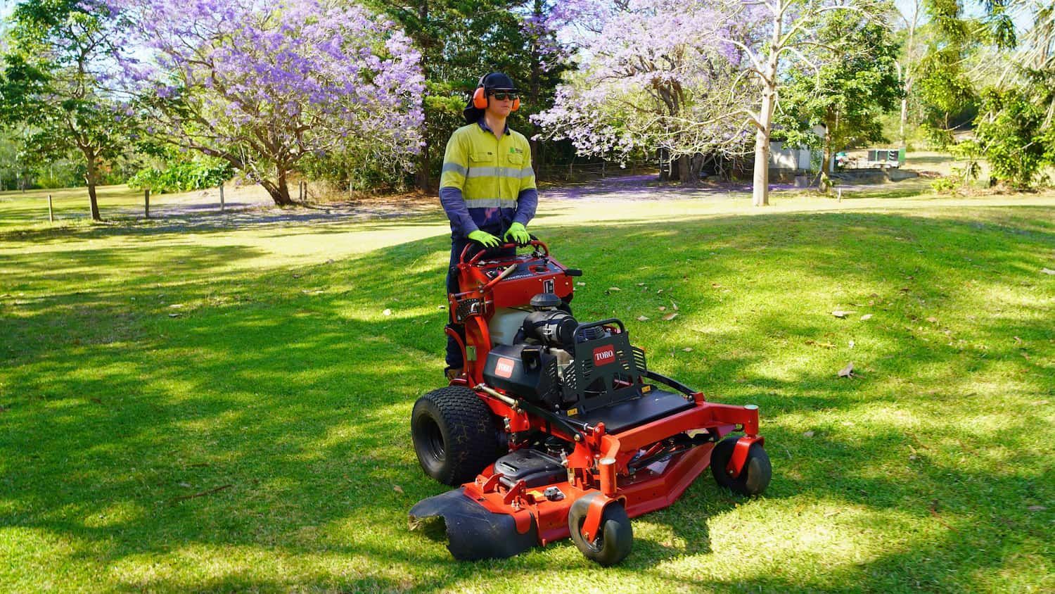 Person operating a red zero-turn mower on a grassy lawn. Purple flowering trees in the background.