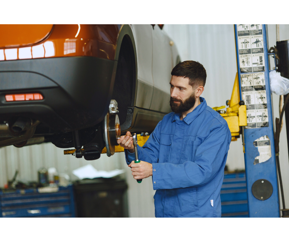 Mechanic in blue overalls, working on a car's brakes, which is raised on a lift in a garage.