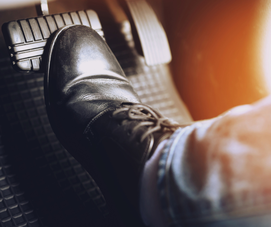Foot in a black shoe pressing down on a car's brake pedal.