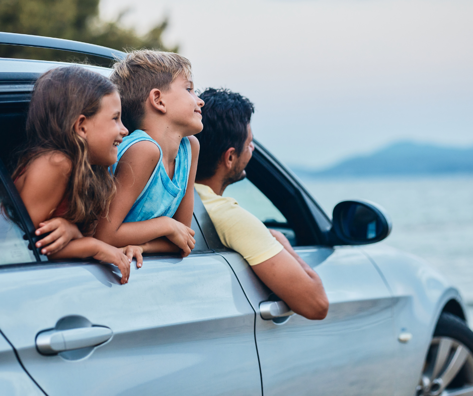 A family in a car, looking out at the ocean. A girl, boy, and man gaze towards the water.