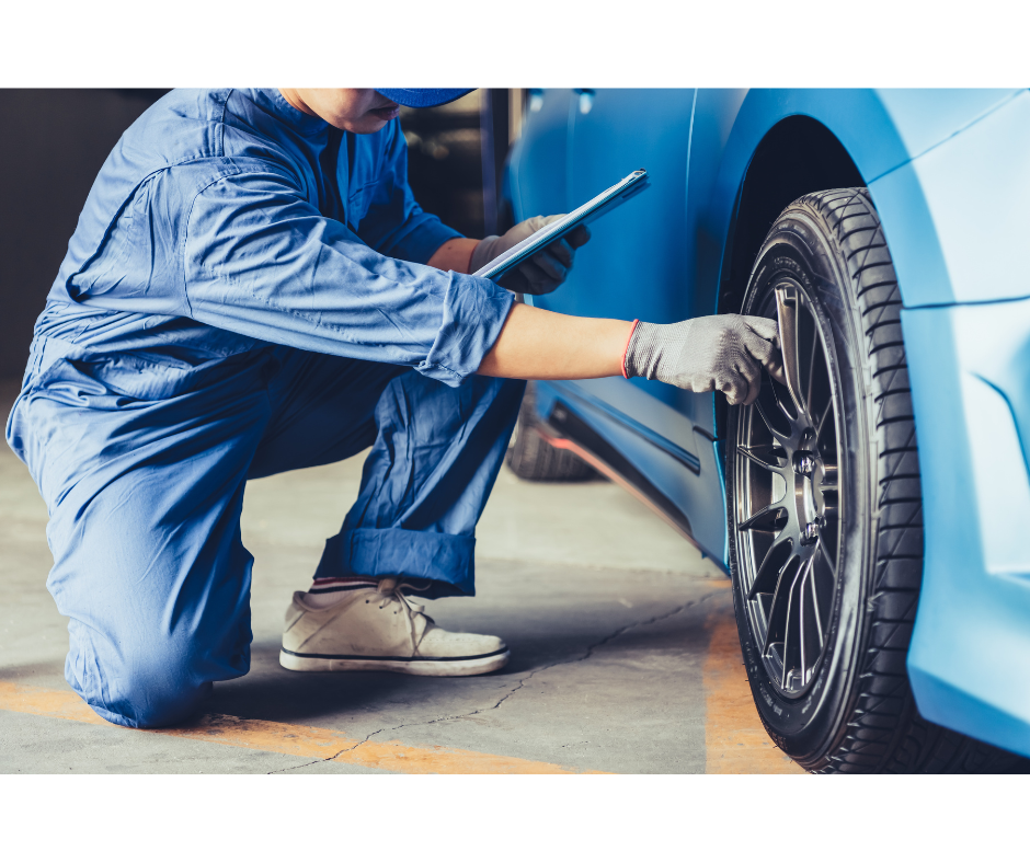 Mechanic in blue overalls inspecting a blue car's tire, holding a clipboard, inside a garage.