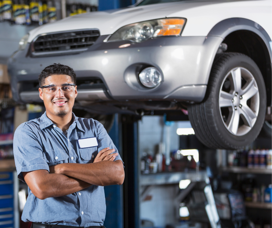 Mechanic with safety glasses smiles, arms crossed, in front of a car raised on a lift in a garage.