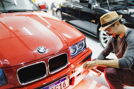 Man inspecting a red BMW in a garage, wearing a hat and glasses. Another black car is visible in the background.