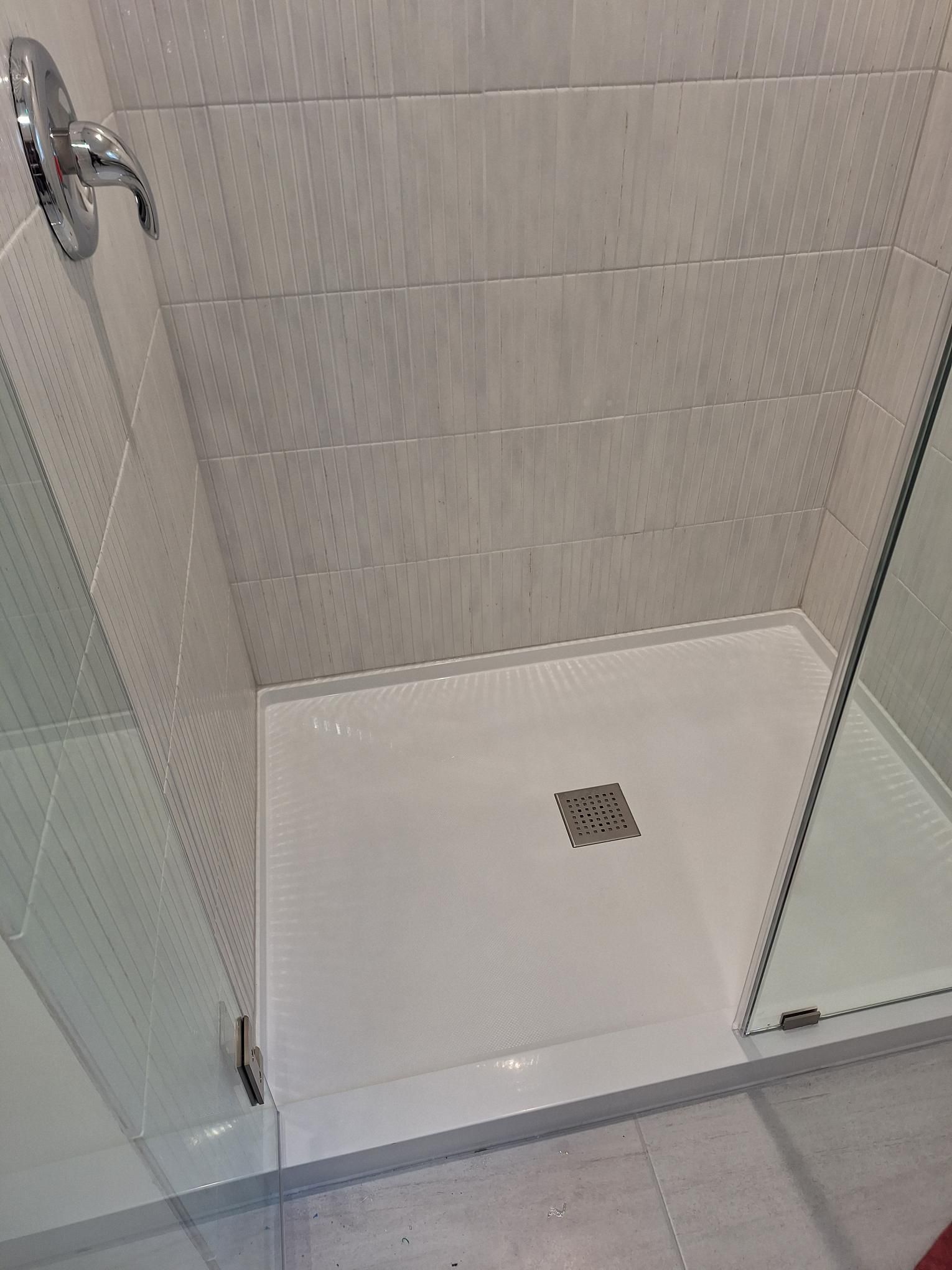 Man Using a Grey Vacuum Shower with white tiles, a glass door, and a white shower base with a drain.