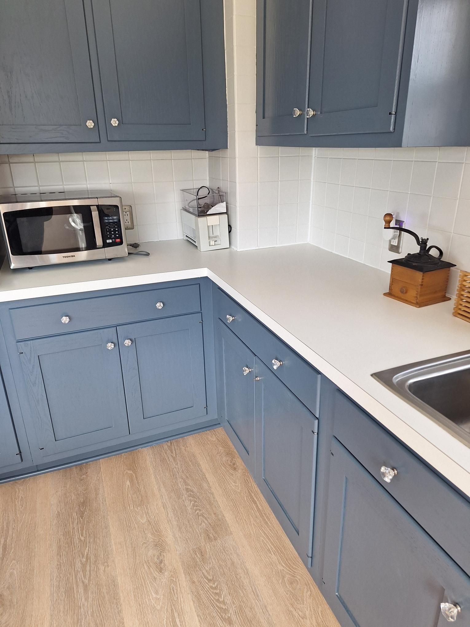 Blue kitchen cabinets and white countertop, with microwave, coffee grinder, and sink.