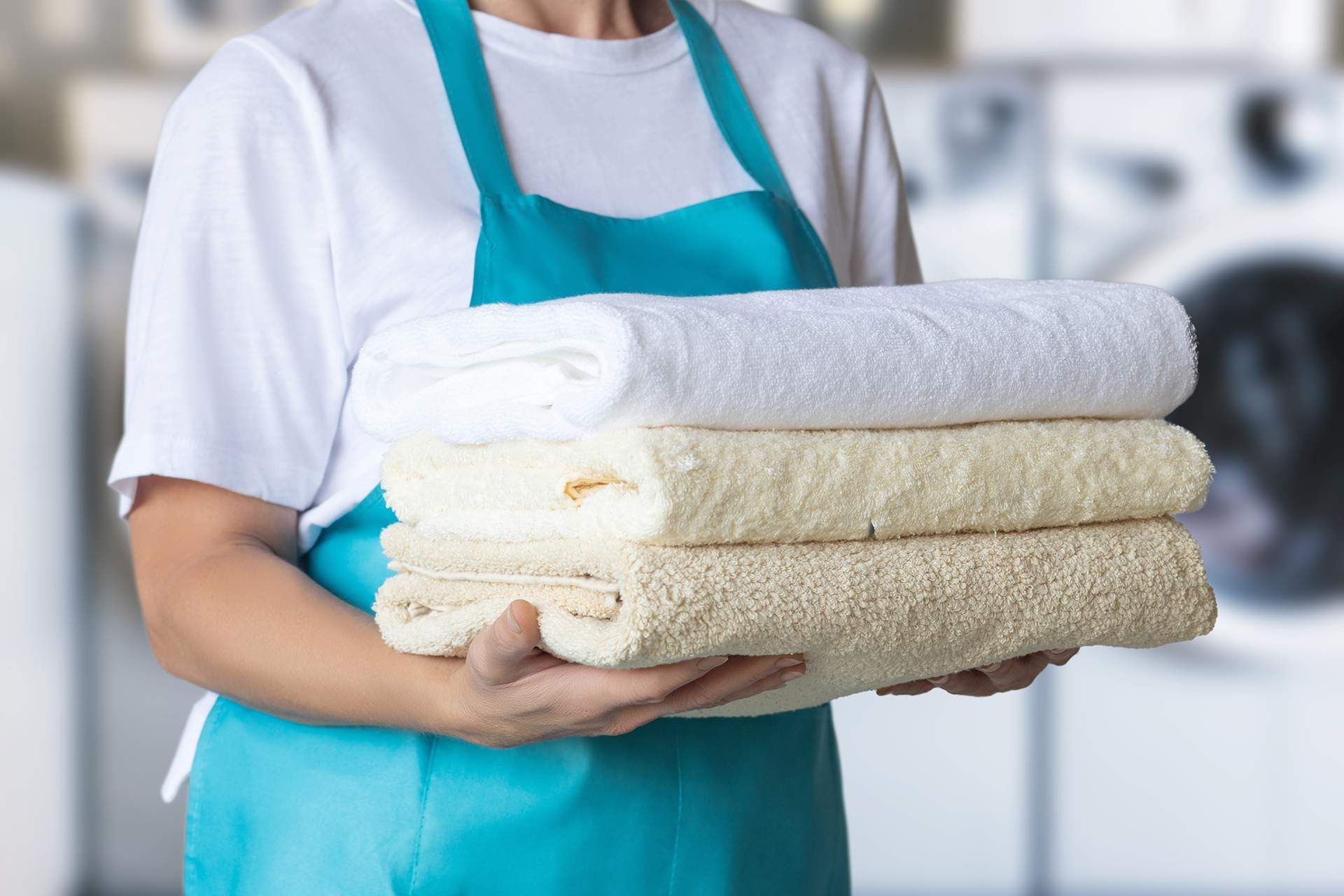 Woman in turquoise apron holding a stack of folded white and beige towels in a laundromat.