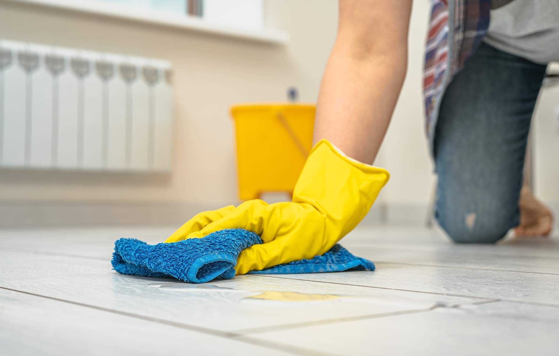 Person wearing yellow gloves, cleaning white tiled floor with a blue cloth; a yellow bucket is visible.