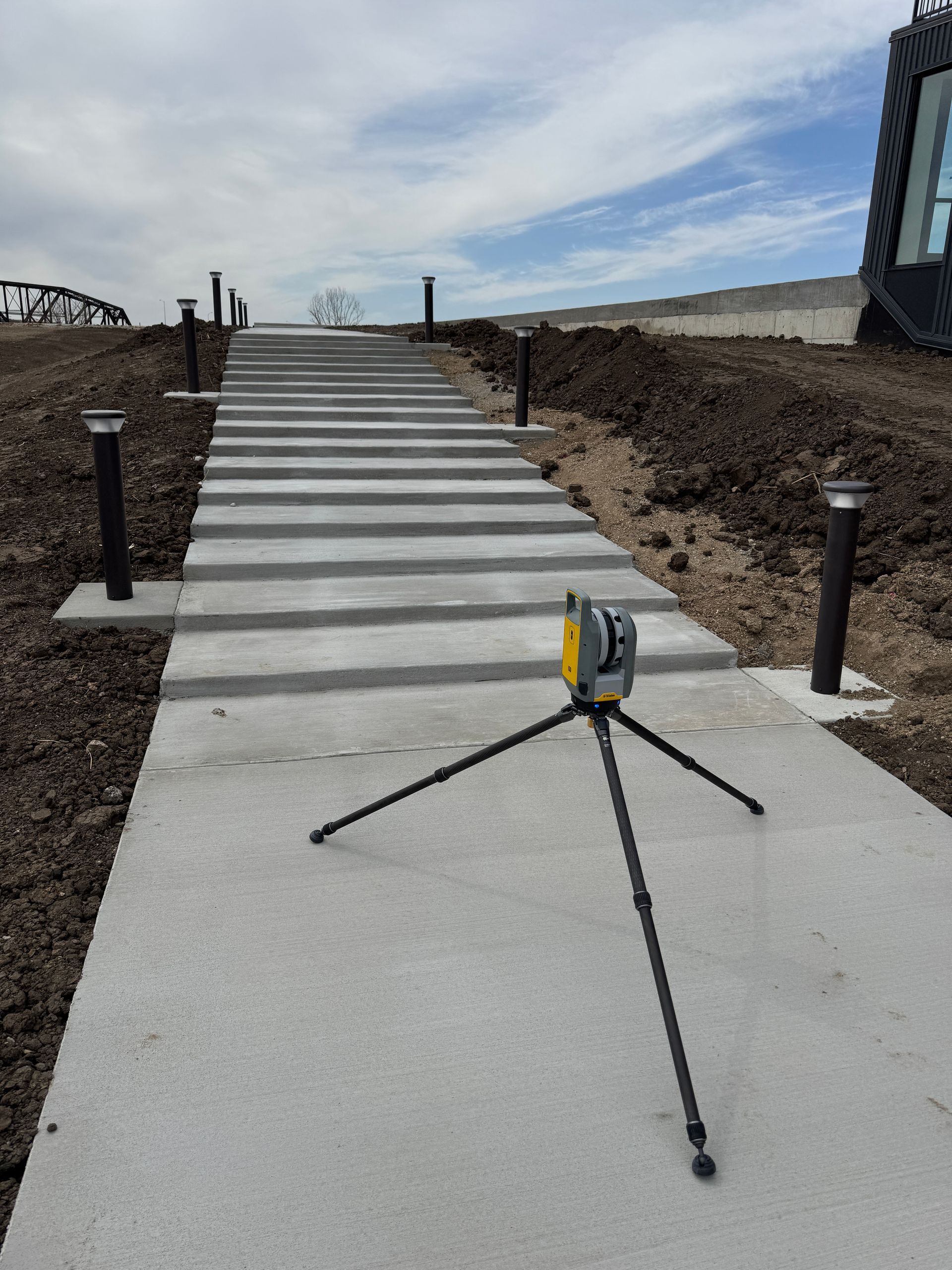 A tripod for laser scanning is sitting on a sidewalk next to a set of stairs 