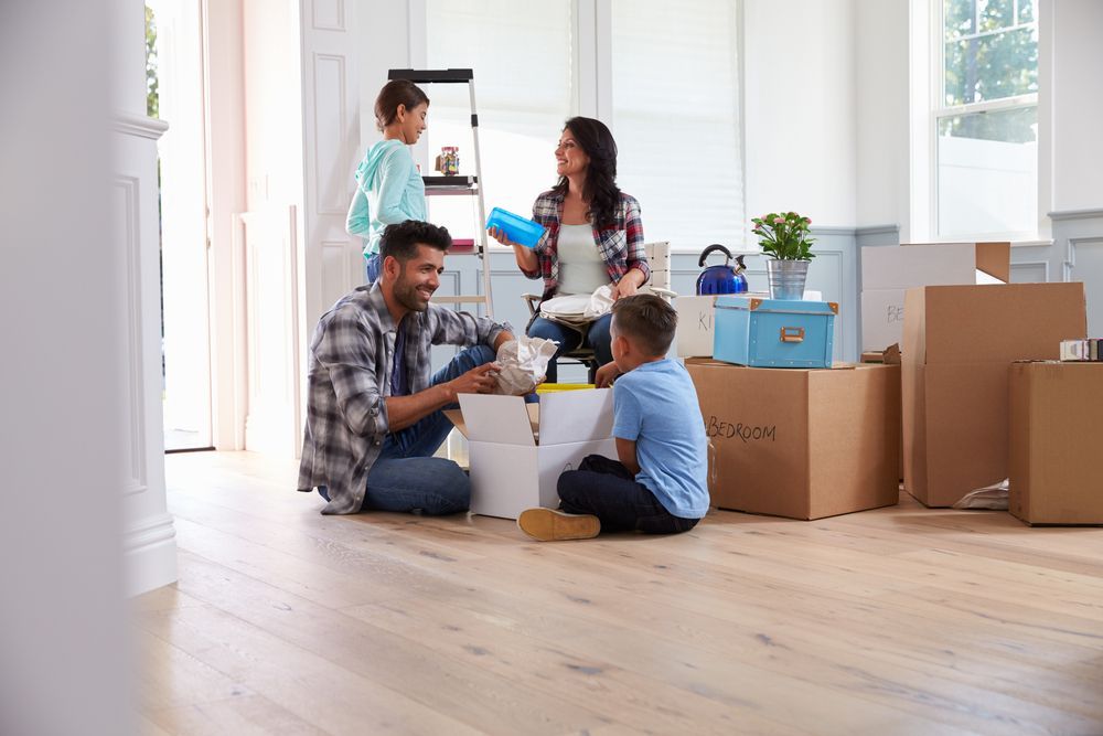 A family is sitting on the floor in a living room surrounded by moving boxes.