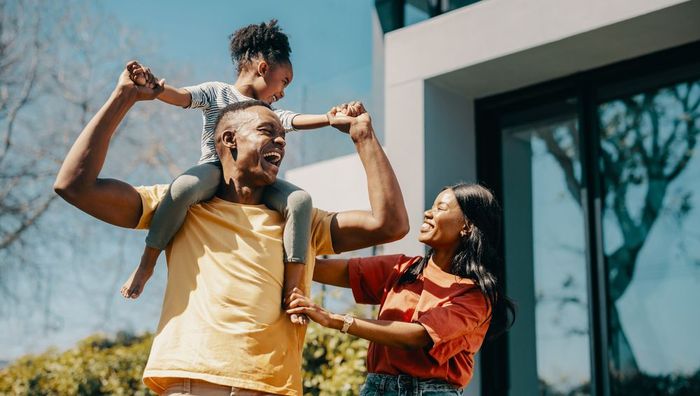 A man is carrying a child on his shoulders in front of a house.