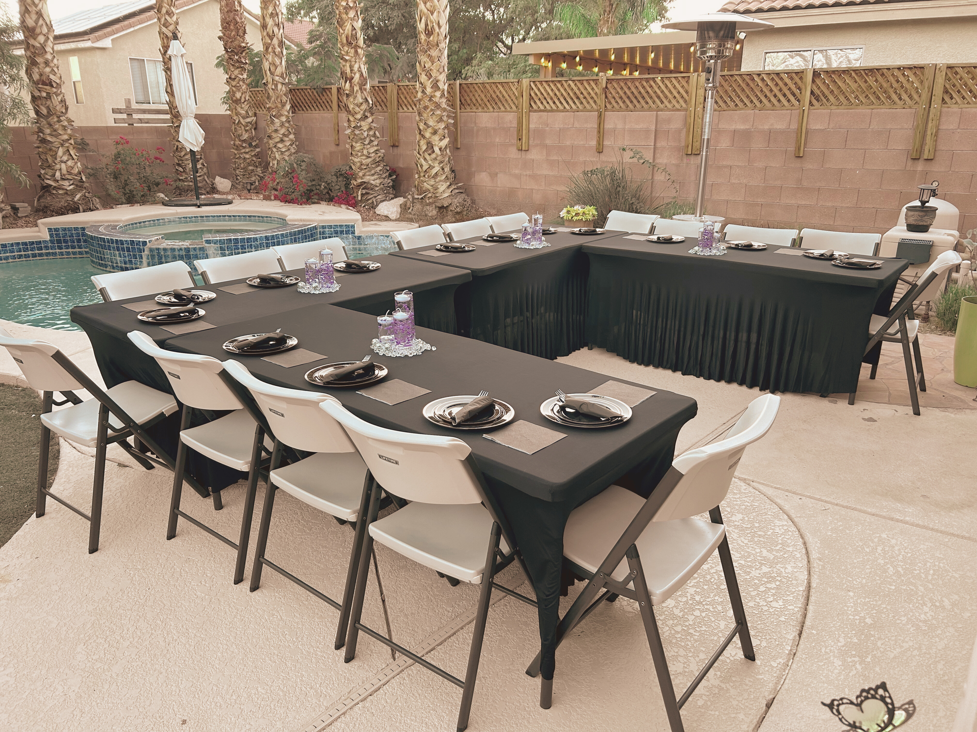 Outdoor party setup with a U-shaped table covered in black linens. White chairs surround the table near a pool.