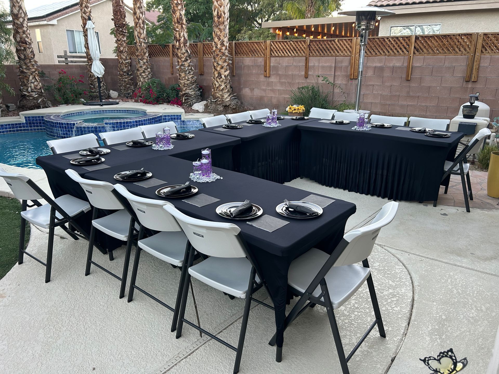 Outdoor party setup with a U-shaped table covered in black linens. White chairs surround the table near a pool.