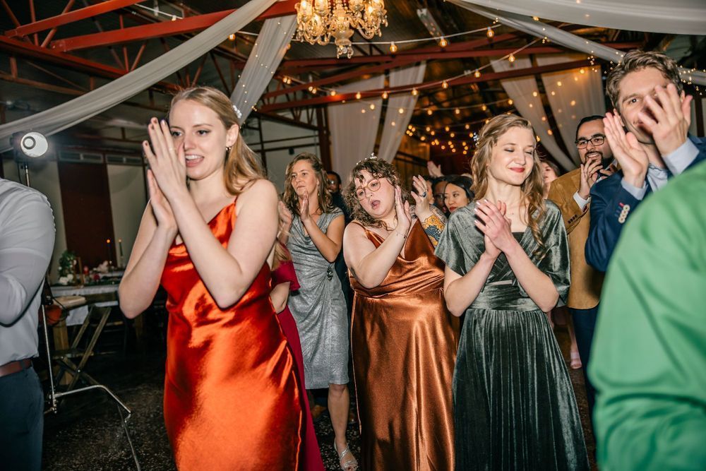 People clapping at an indoor celebration. Women in formal gowns smile, illuminated by string lights.