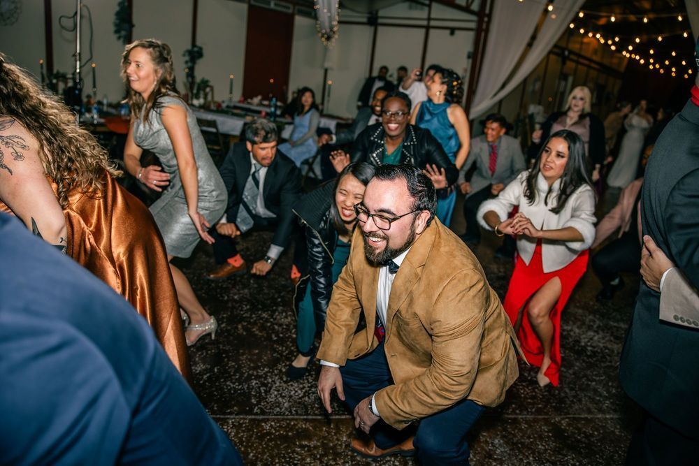 People dancing energetically at a wedding reception. Some are crouching, others smiling, under lights.