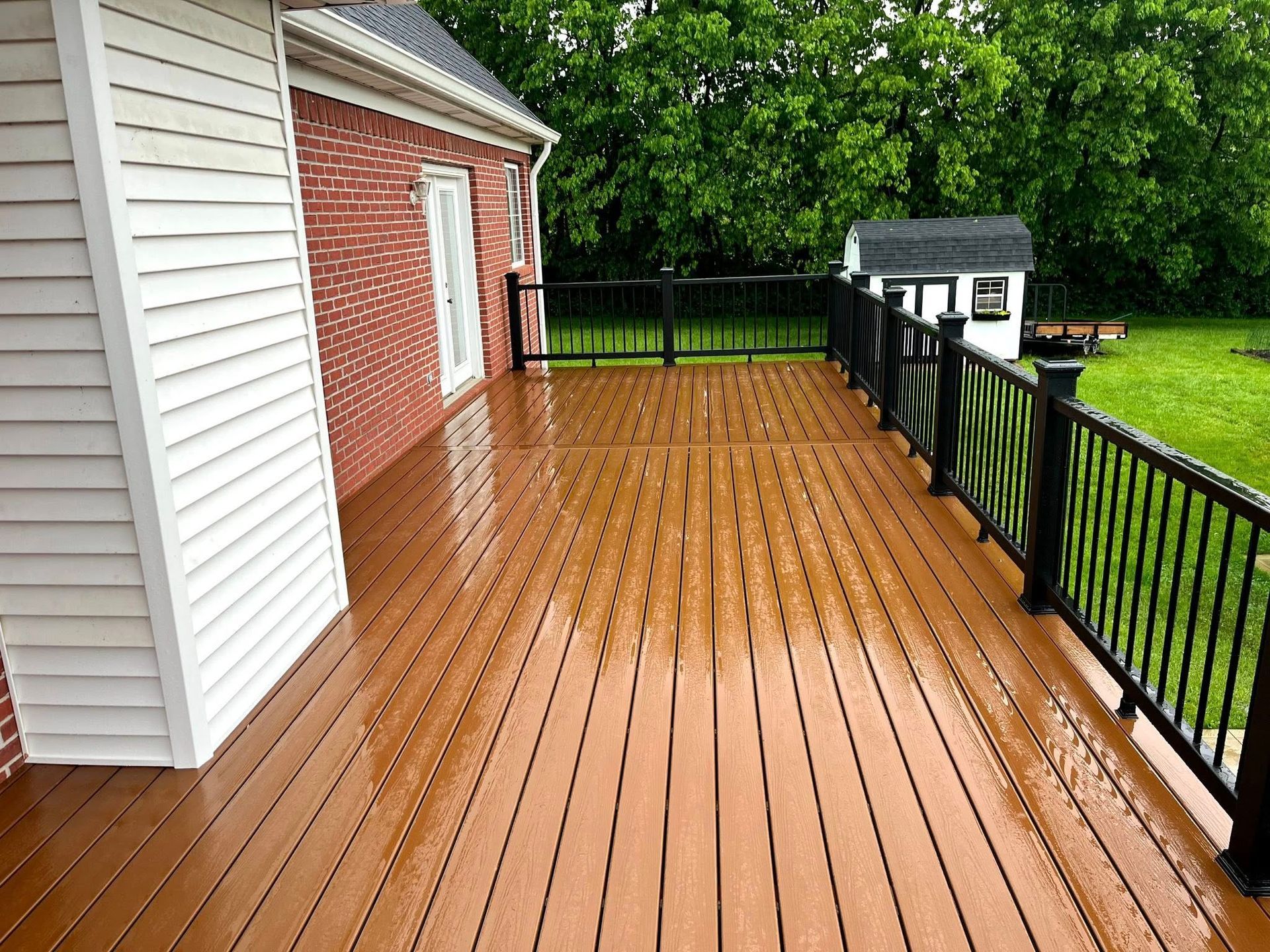 A large wooden deck with a black railing and a brick house in the background.