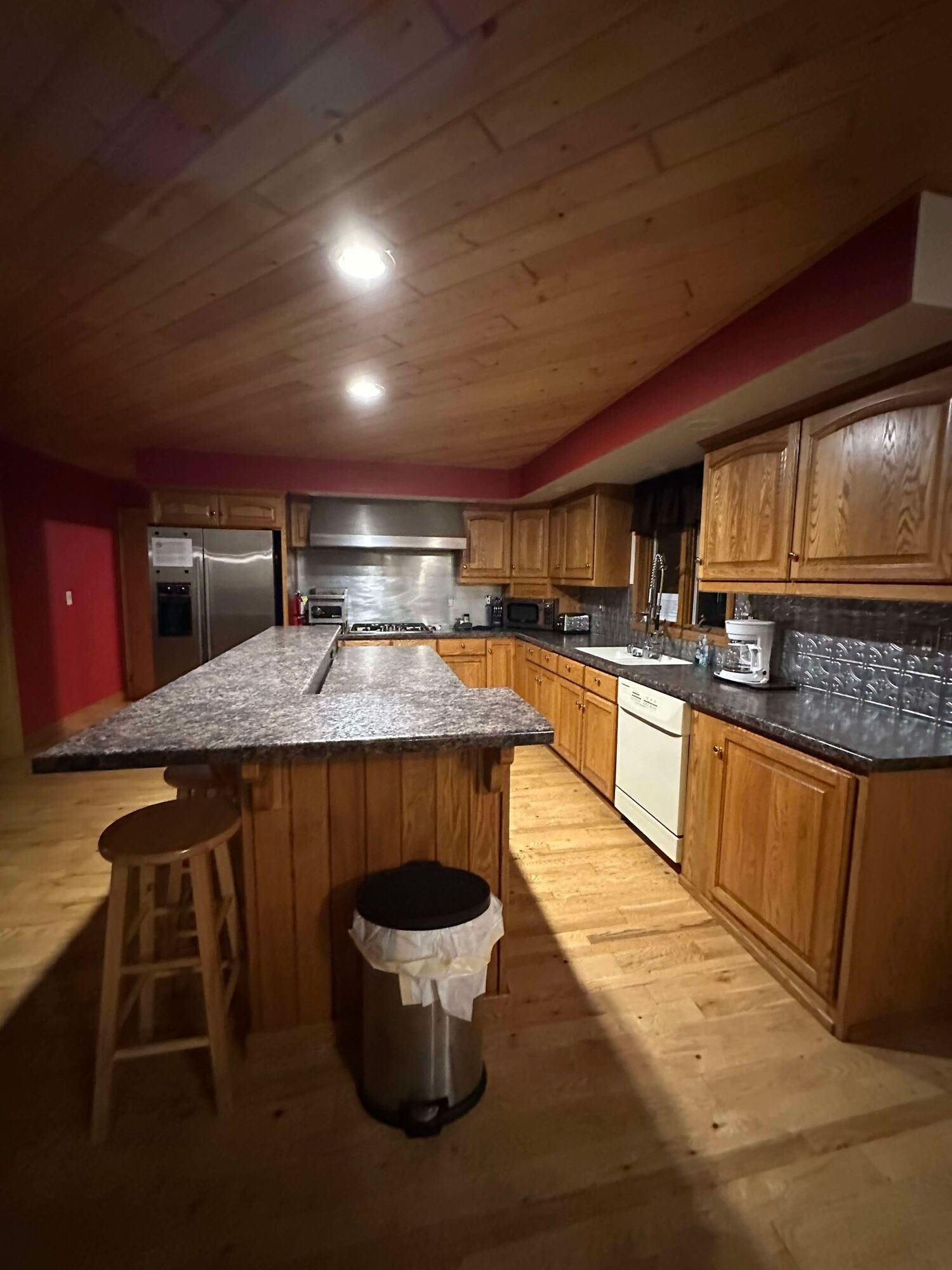A kitchen with wooden cabinets , granite counter tops , stools and a trash can.