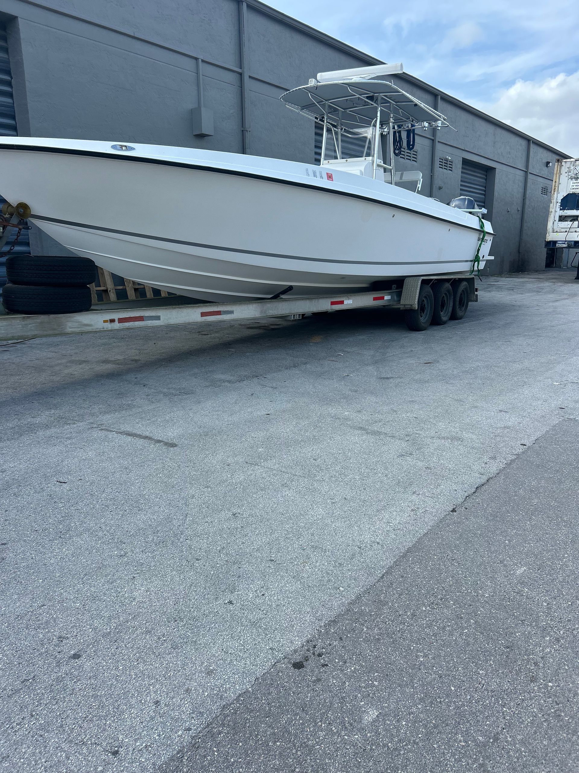 White boat on a trailer in front of a gray building. Cloudy sky above.