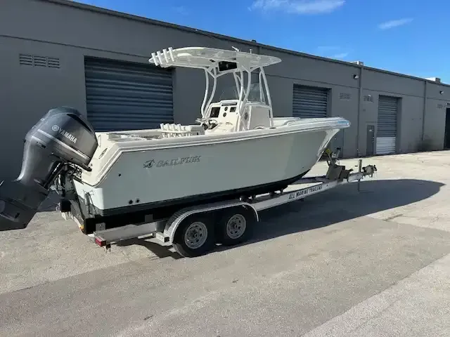 A light-colored Sailfish boat on a trailer in front of a gray industrial building on a sunny day.
