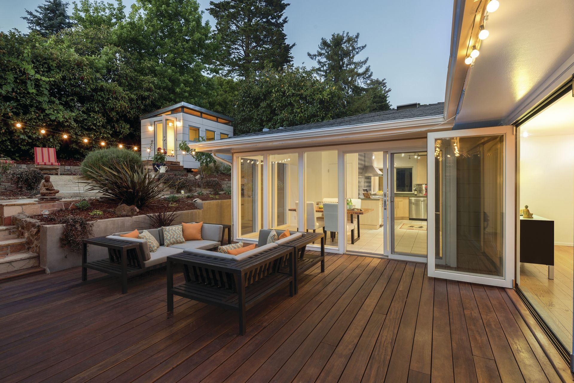 Wooden deck with outdoor seating, opening to a kitchen, lit by string lights and surrounded by greenery.